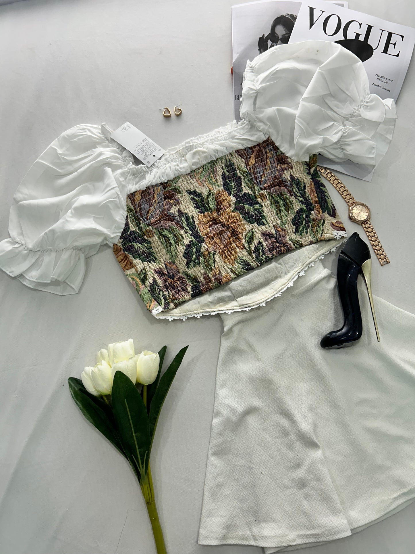 White blouse with ruffled details, floral-patterned skirt, high-heeled shoes, white tulips, and a magazine on a light gray surface.